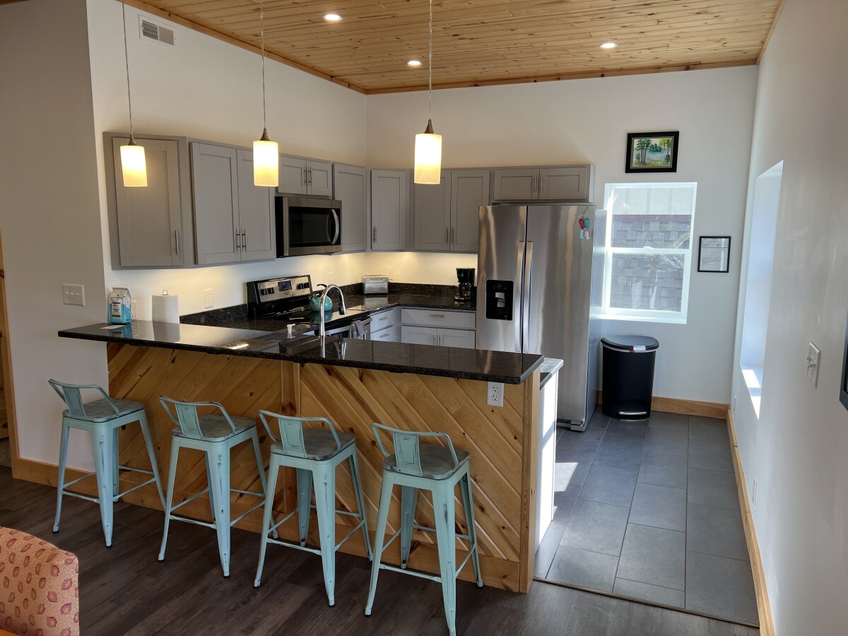 A modern kitchen is showcased, featuring a large island with four metal stools. Gray cabinetry complements black countertops while stainless steel appliances are included. Natural light filters in through a nearby window, illuminating the tiled flooring and creating a warm atmosphere.