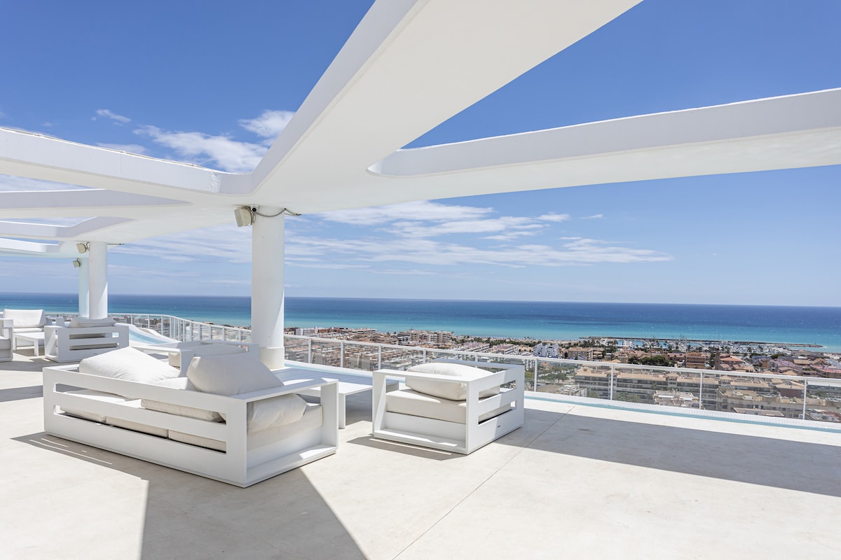 A spacious rooftop terrace features modern white lounge furniture arranged for relaxation. The expansive view showcases the coastline and sea under a clear blue sky, with sunlight illuminating the area. The open layout provides an inviting space for enjoying outdoor moments.