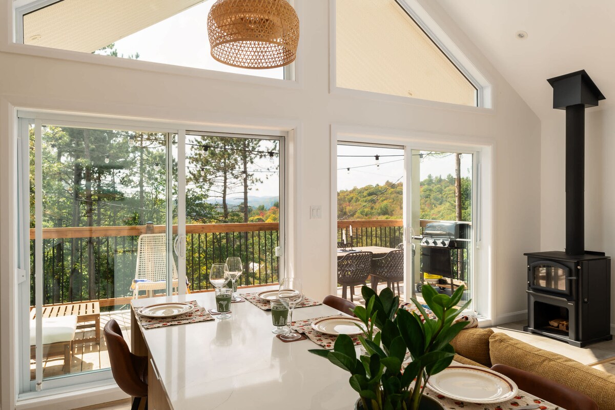 An open-plan dining area features a large island table, placed opposite expansive glass doors that provide views of the surrounding nature. A wood-burning stove is situated to one side, while a cozy seating area is visible nearby. Soft natural light brightens the space.