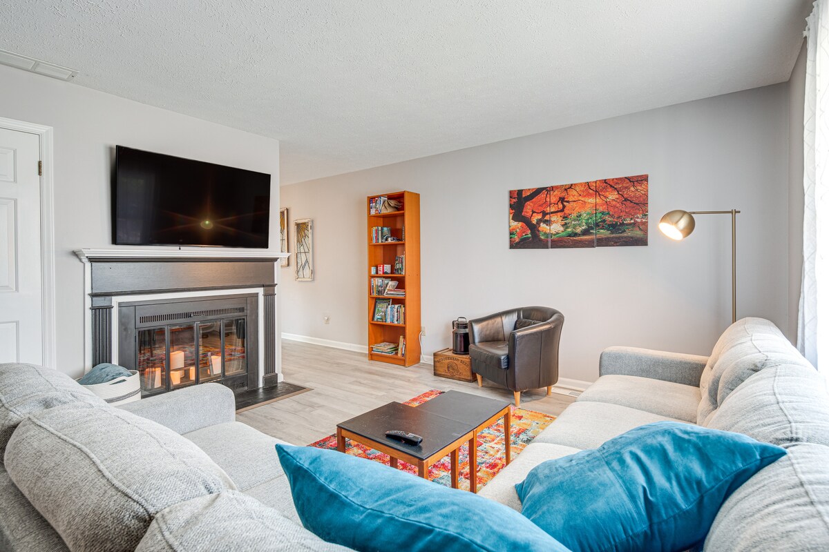 A spacious living area is highlighted by a large sectional sofa with blue accent pillows. A flat-screen TV is mounted above a decorative fireplace. A bookshelf and a vibrant artwork depict a natural scene, contributing to a casual atmosphere.