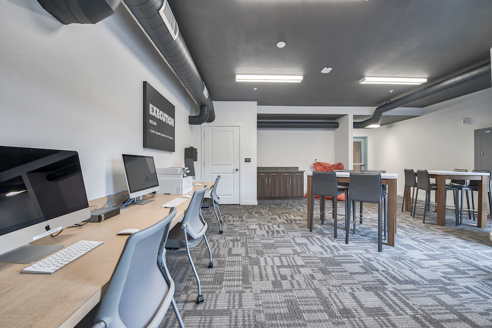 A communal workspace is displayed, featuring multiple wooden desks with computers and ergonomic chairs. Soft lighting is provided by overhead fixtures, and gray carpeting enhances the modern industrial look of the area. A lounge area is visible in the background, with additional seating options.