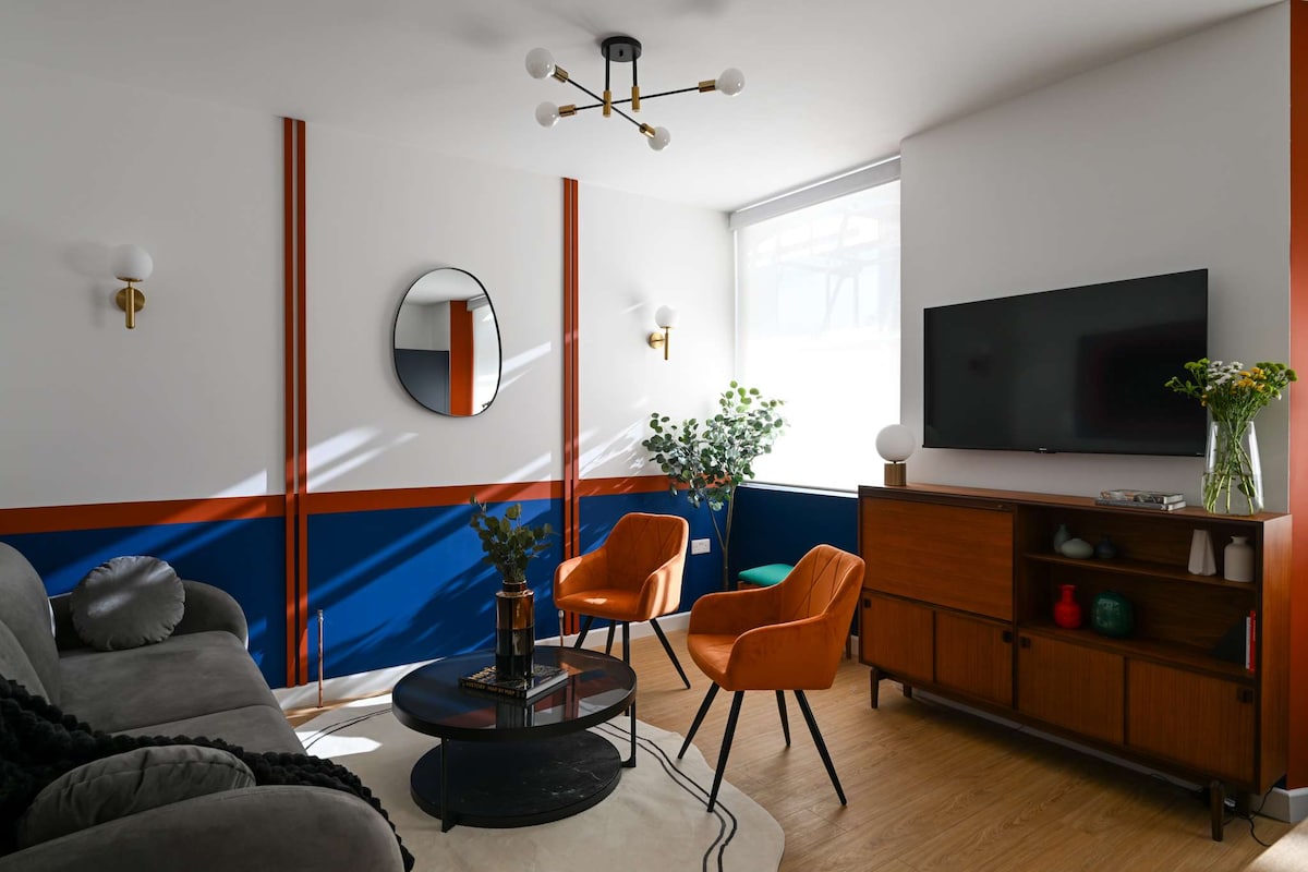 A modern living space features a stylish gray sofa and two orange accent chairs. Natural light fills the room through a large window, highlighting the blue and white walls. A round coffee table sits centrally, complemented by a mid-century storage console and decorative plants.