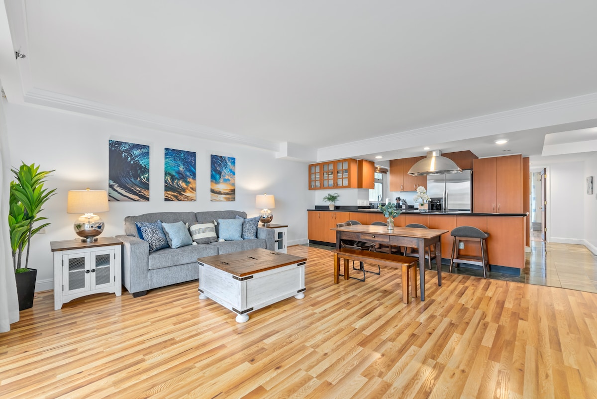 A spacious living area features a comfortable gray sofa with decorative pillows, accompanied by a wooden coffee table. Two pieces of artwork hang on the wall above, and the kitchen is located in the background, showcasing modern cabinetry and appliances. Natural light fills the room, enhancing the hardwood flooring.