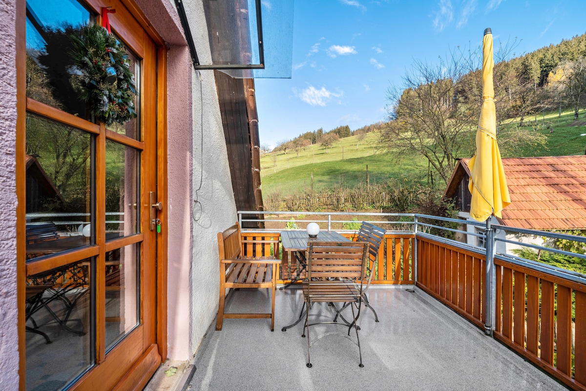 A private balcony features a wooden table and chairs, offering a view of rolling hills and greenery. A yellow umbrella provides shade, while a decorated door leads inside. The surface is smooth, contributing to a comfortable outdoor space.