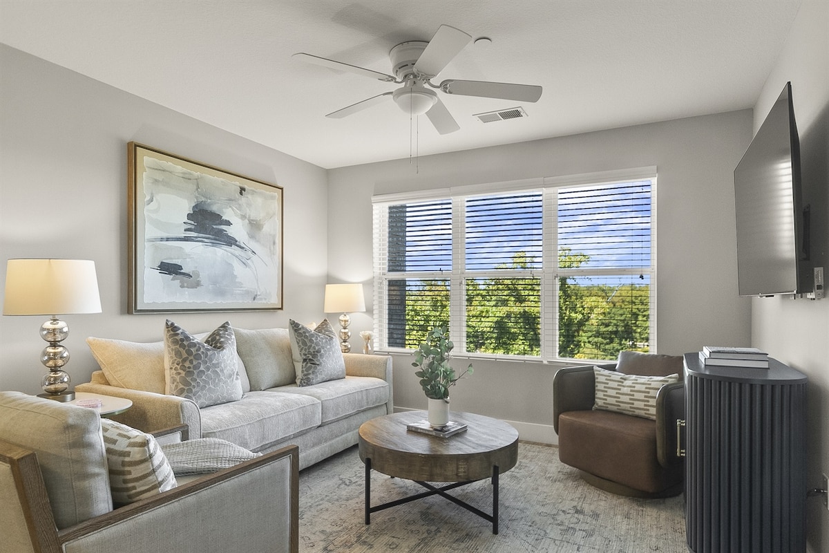 A bright living room features a comfortable sofa and a lounge chair, positioned around a round coffee table. Large windows allow natural light to fill the space, with partially opened blinds revealing a glimpse of greenery outside. Light-colored walls are complemented by decorative artwork.