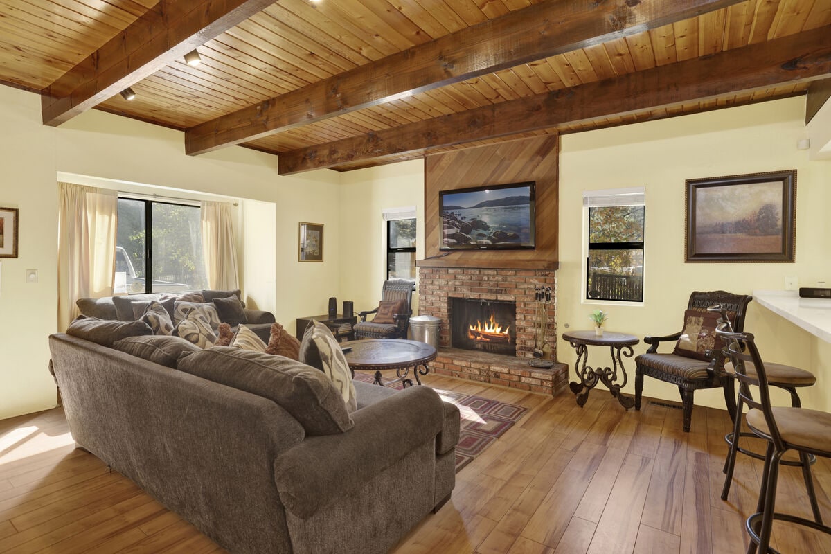 A spacious living area features a cozy seating arrangement with a large sofa and chairs, all oriented towards a brick fireplace. Natural light enters through big windows, and warm wood beams highlight the ceiling. Decorative artwork adds a touch of character to the walls.