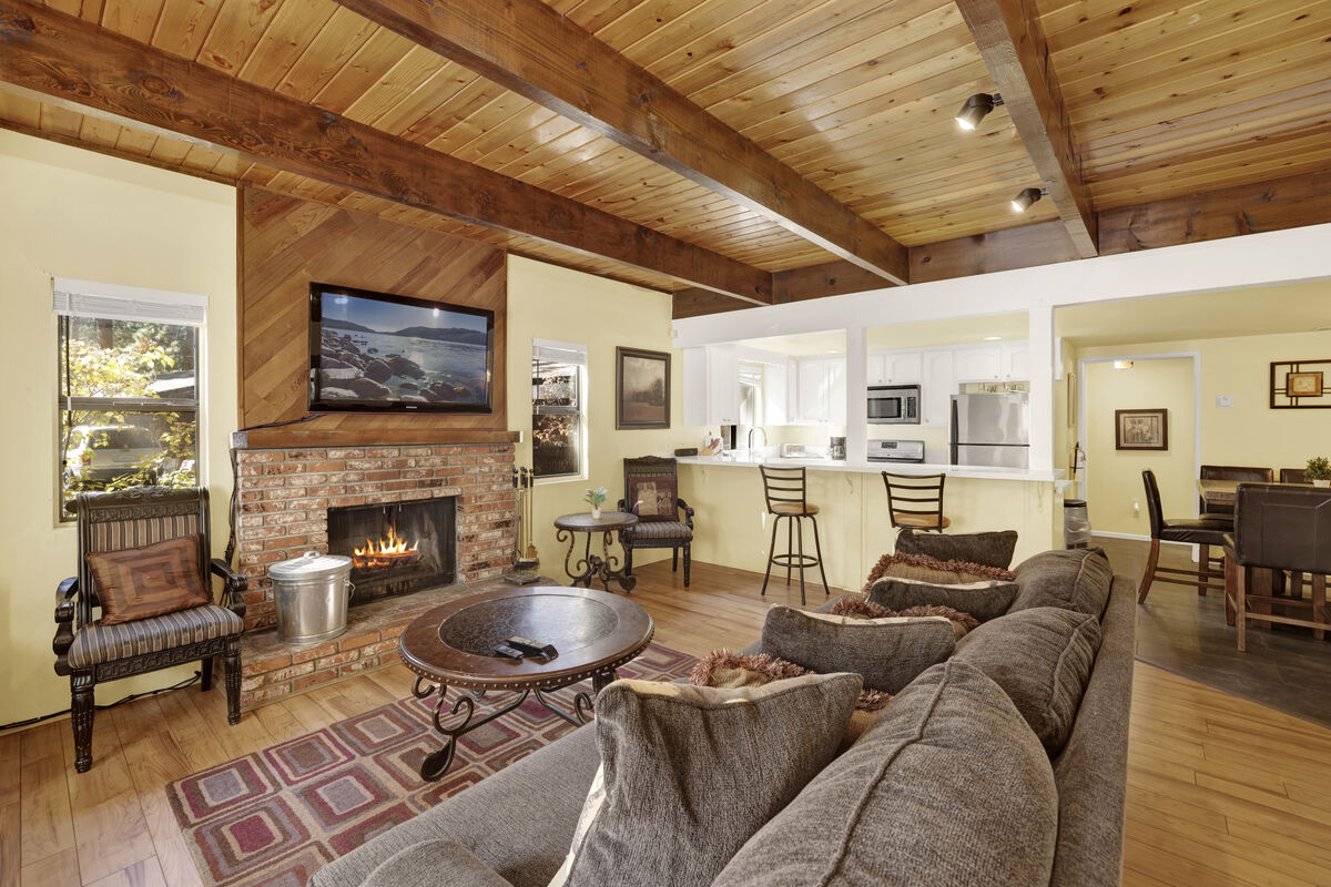 A spacious living area features a comfortable sectional sofa arranged around a low coffee table. A stone fireplace with a TV mounted above serves as a focal point. The wood-beamed ceiling and warm yellow walls add to the inviting ambiance. A bar area with stools can be seen in the background.