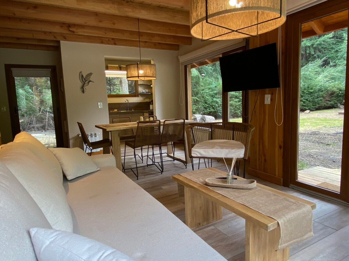 A cozy living area features a light-colored sofa and wooden coffee table. Dining options are available with a nearby table and chairs. Large glass doors provide natural light and views of the surrounding forest, while overhead light fixtures enhance the ambiance.