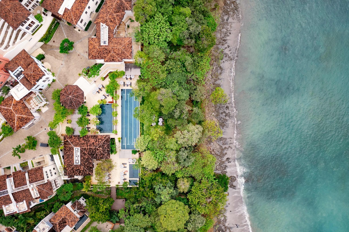 An aerial view captures the layout of Casa Maya and its lush surroundings, highlighting the proximity to a sandy beach. The property is bordered by greenery, with a communal pool visible among the buildings, while the ocean exhibits shades of blue near the shoreline.