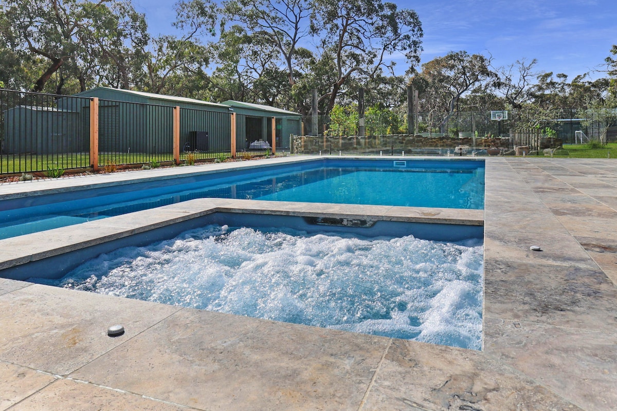 A spacious outdoor area features a heated pool and attached spa, both surrounded by a natural landscape of trees. The poolside area is paved with light-colored stone, and a fence provides privacy. Gentle water movement creates a calming atmosphere.