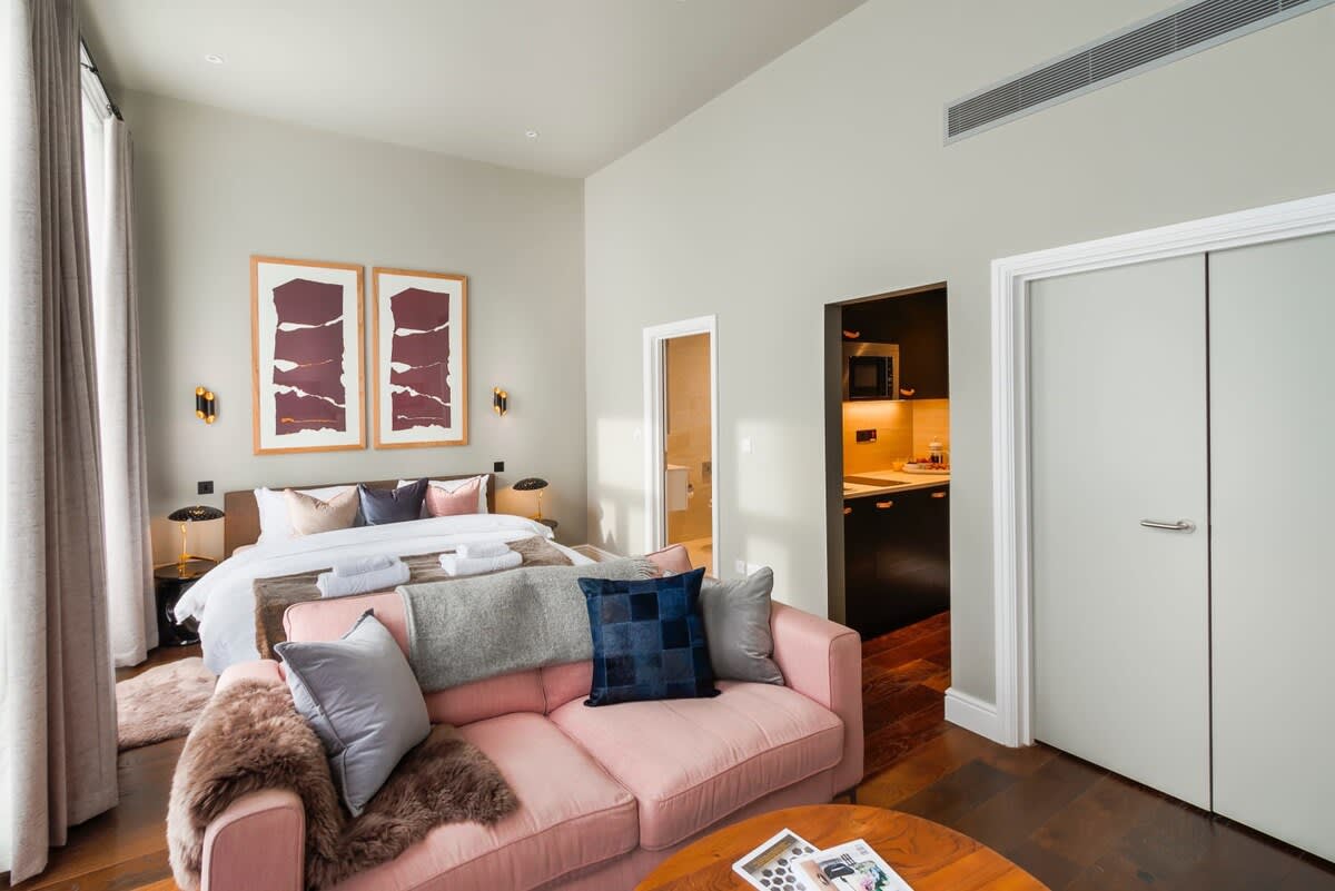 A spacious studio is depicted, featuring a king-size bed with crisp linens in the background. A comfortable pink sofa is positioned in the foreground, adorned with decorative pillows and a soft throw. An open doorway reveals a kitchenette area, enhancing the studio's functionality.