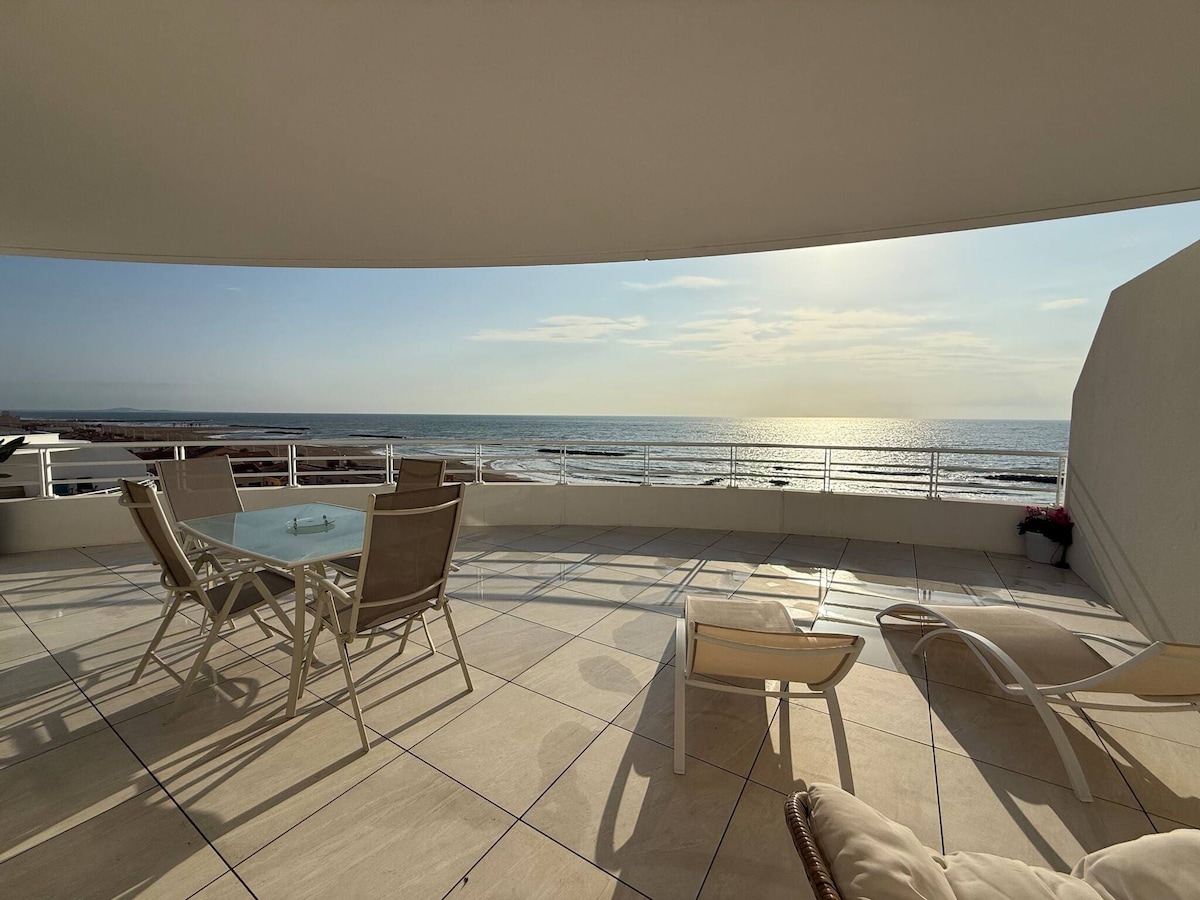 A spacious terrace overlooks the sea, featuring a glass table surrounded by several chairs. Lounge chairs are positioned for sunbathing, and the calm waves can be seen in the distance under a clear sky.