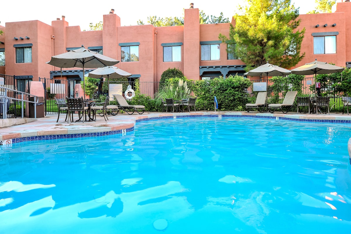 A clear swimming pool reflects the blue sky, surrounded by lounge chairs and umbrellas. Lush greenery and the property buildings are visible in the background, offering a serene setting for relaxation.