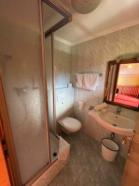 The bathroom features a compact design with a shower enclosed in clear glass. A modern toilet is positioned beside a circular sink mounted on a vanity. Light-colored tiles cover the walls, and fresh towels are neatly arranged on a rack.