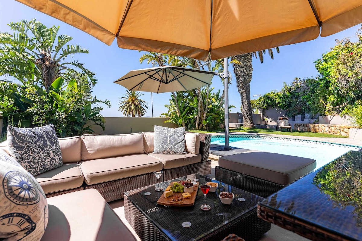 A spacious outdoor lounge area features a comfortable sectional sofa with decorative cushions, positioned under a large umbrella. A coffee table displays an elegant spread of refreshments, while a sparkling pool and lush greenery provide a refreshing backdrop.