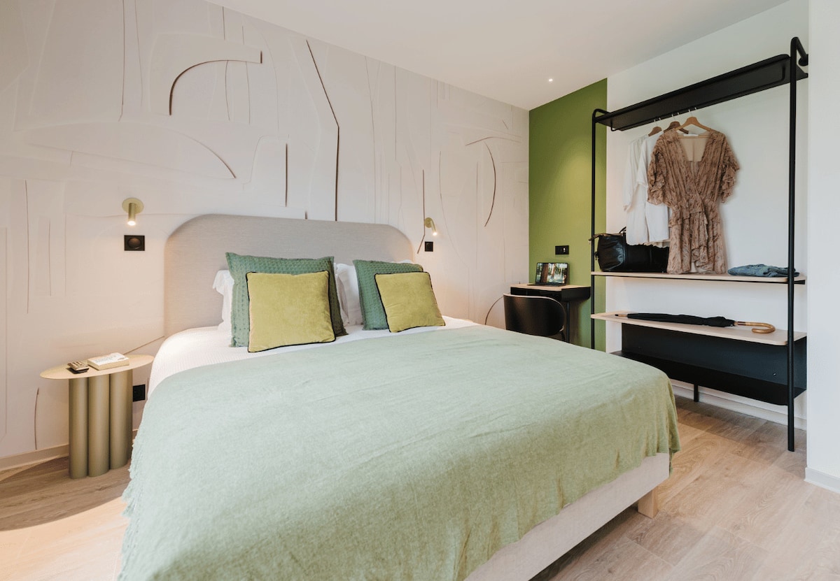 A comfortable bedroom features a spacious bed dressed in soft linens. Green decorative pillows add a pop of color against the neutral wall with textured designs. A minimalist wardrobe and storage area is visible, showcasing neatly hung clothing, while a small bedside table holds essentials.