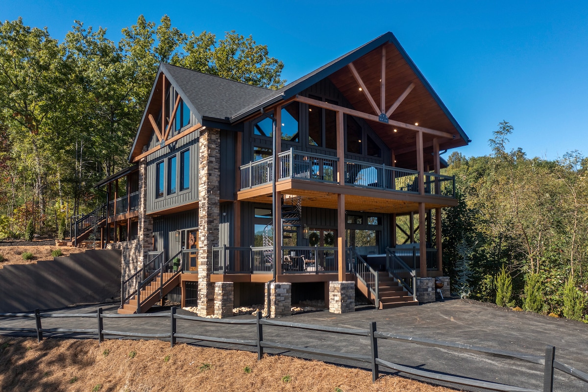The exterior of the cabin showcases a two-story design with a combination of wood and stone features. A large wrap-around deck is accentuated by black railings, offering ample outdoor seating. Lush greenery surrounds the property, enhancing its serene mountain setting.
