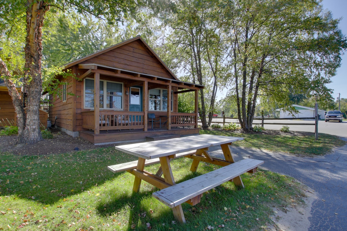 A charming cabin is framed by tall trees, featuring a covered porch with bench seating. A picnic table is positioned in front, ideal for outdoor meals. The surrounding grassy area adds to the inviting appearance of the space.