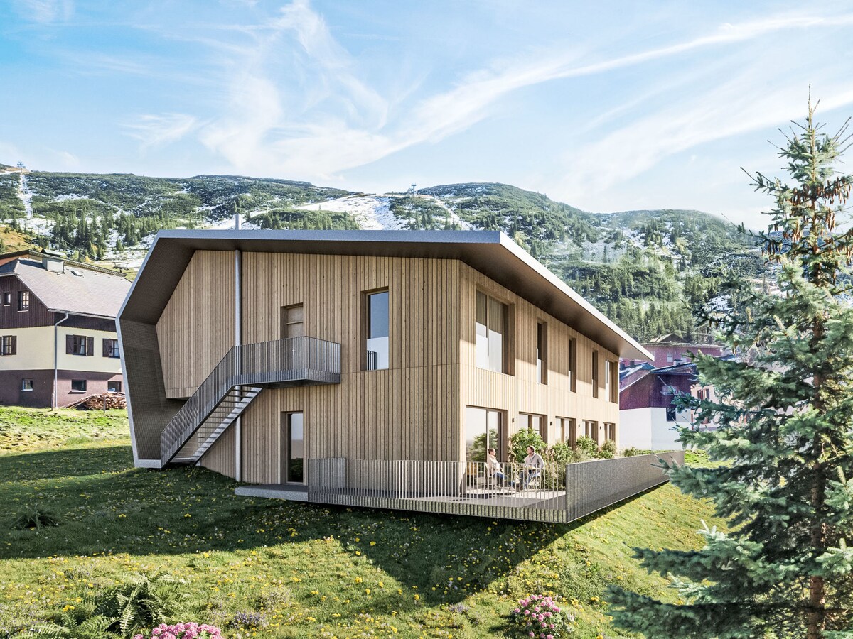 The modern apartment block features a unique architectural design with wooden siding and multiple large windows. The building is situated on a grassy hillside, surrounded by lush vegetation. A staircase leads to an elevated entrance, while the backdrop reveals distant mountains and ski slopes.