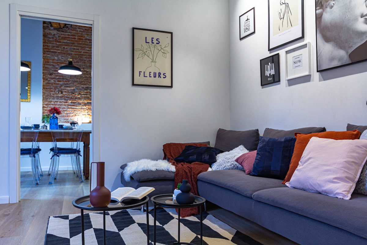 A spacious living area features a comfortable sofa adorned with various cushions in warm hues. Two small coffee tables sit on a patterned rug, alongside a book and decorative items. An open doorway reveals a dining space with a wooden table set for meals.