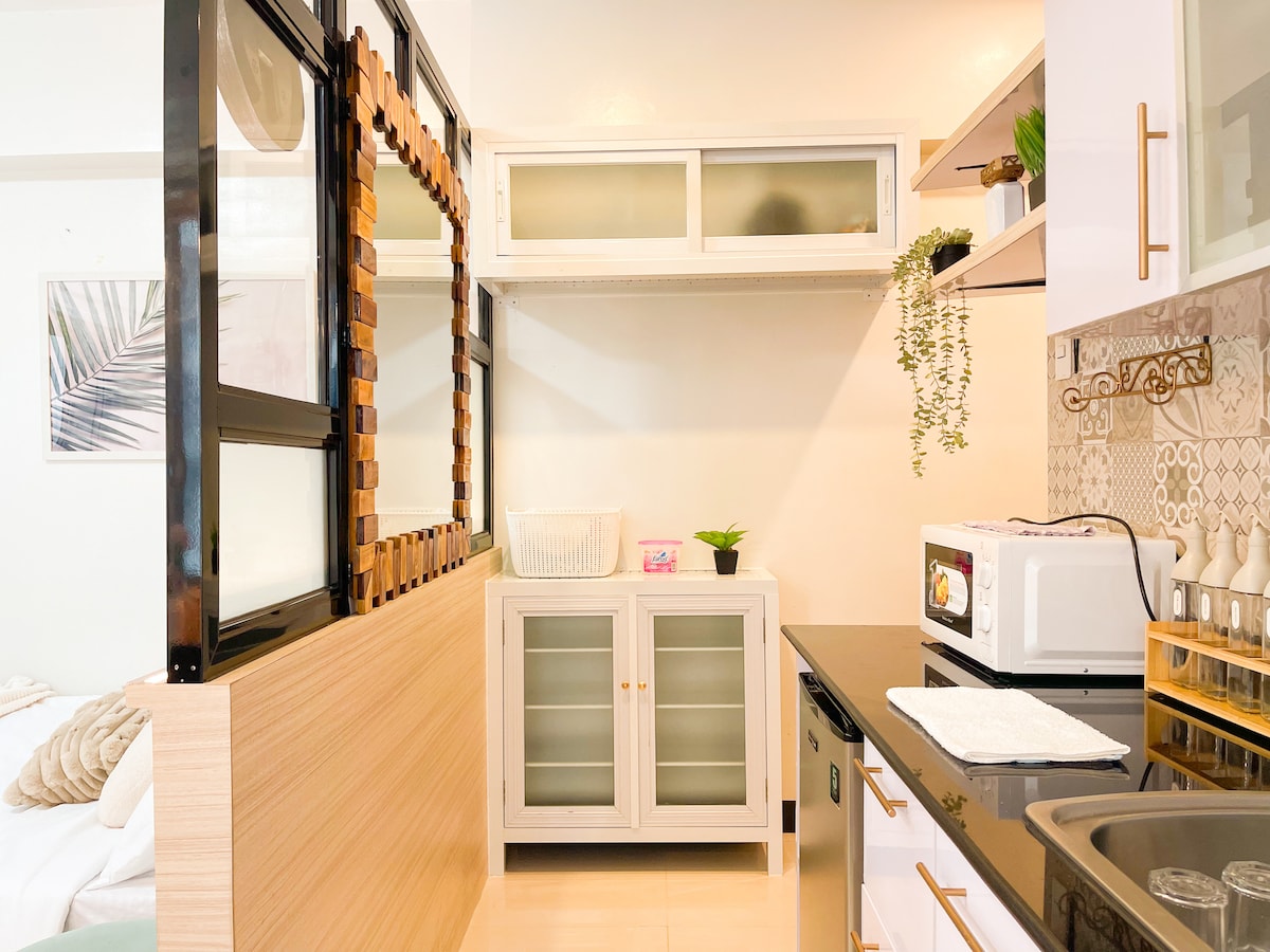 A compact kitchen is shown, featuring modern cabinetry and surfaces. Essential appliances, including a microwave and a refrigerator, are visible. A small plant adds a natural touch, while the countertop is clear and ready for meal preparation. Glass cabinet doors provide storage visibility.