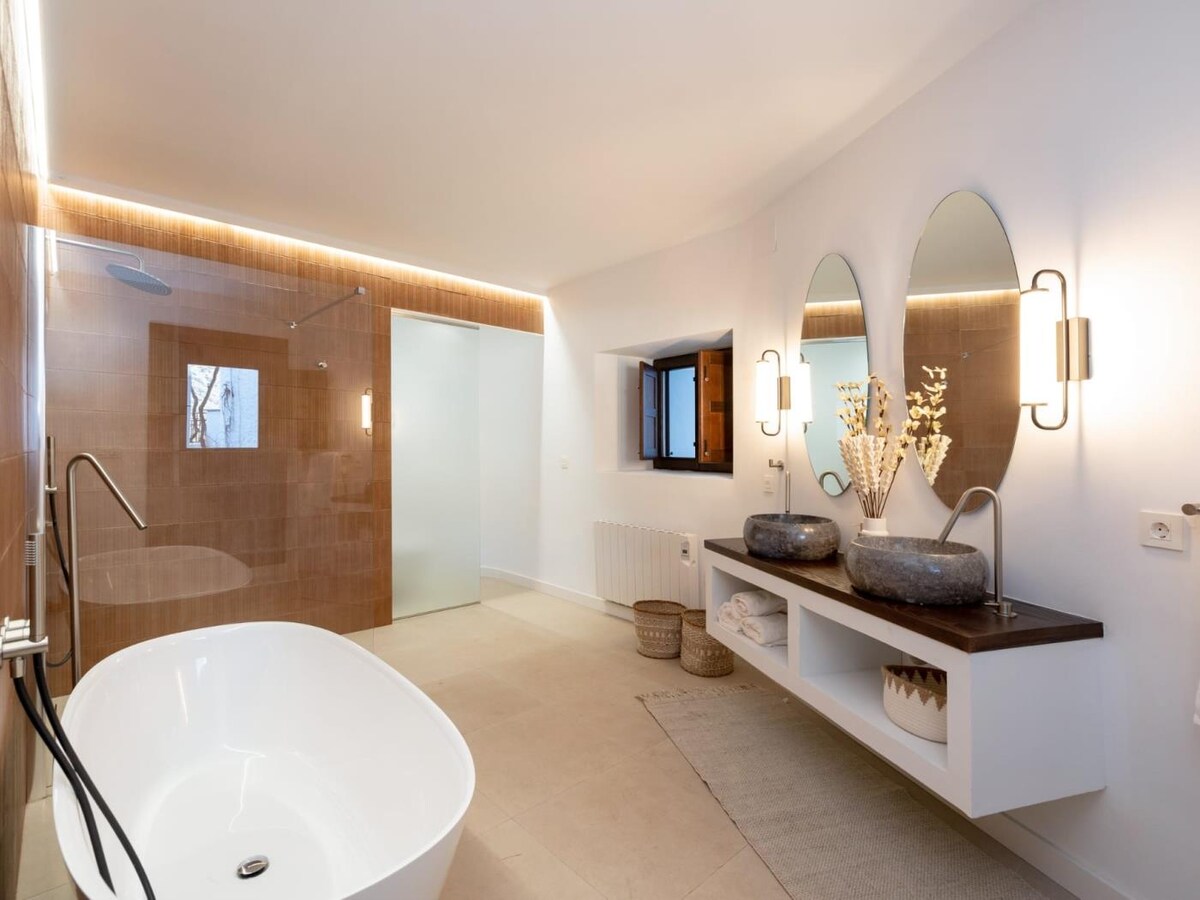 A spacious and well-lit bathroom is presented, featuring a freestanding bathtub and dual sinks with large mirrors. Natural light enters through a small window, illuminating the neutral-colored walls and floor. Shelving below holds neatly stacked towels and decorative baskets.