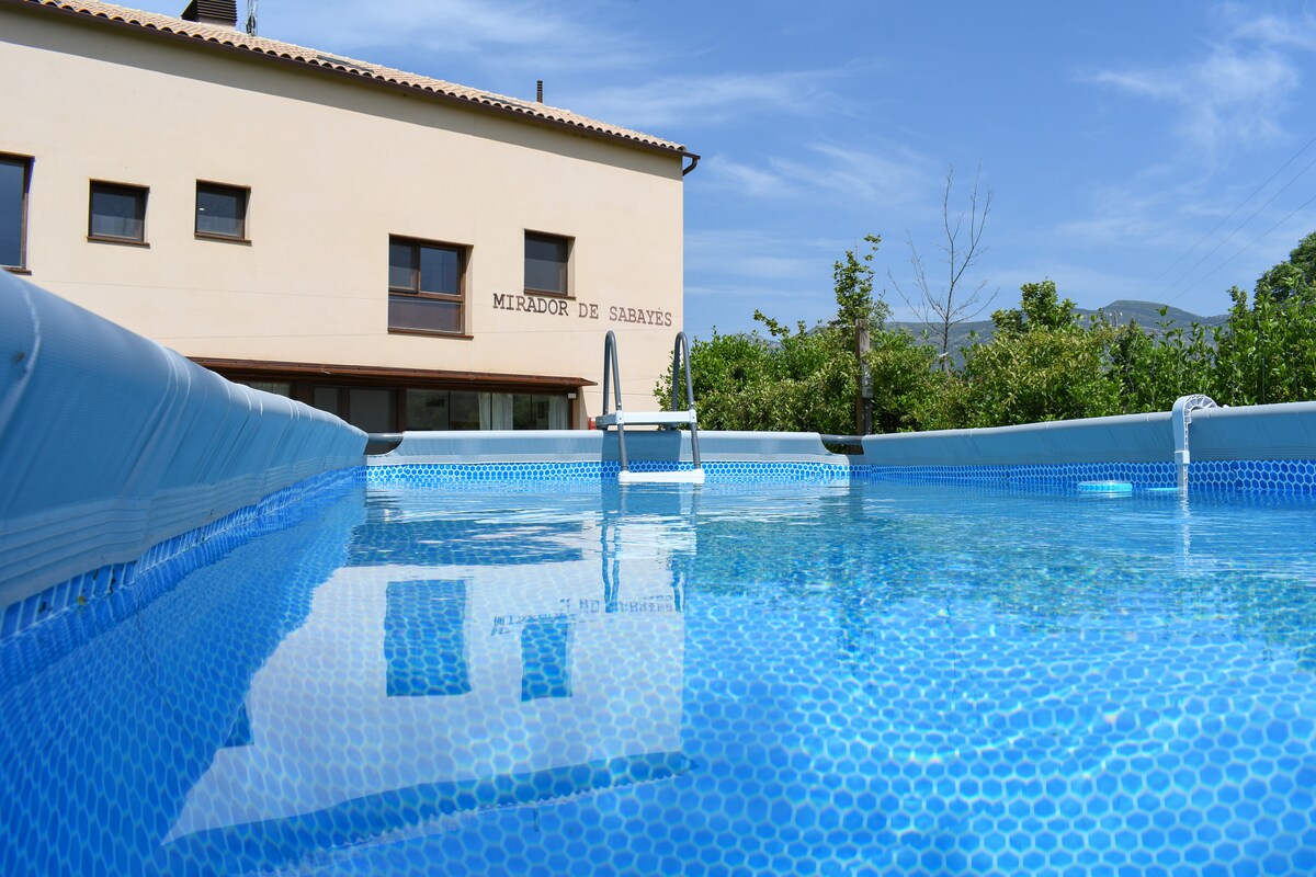 A private swimming pool is featured in the foreground, with clear blue water reflecting the sky. The back of the house, labeled 'Mirador de Sabayés,' is visible, surrounded by greenery and mountains in the distance.