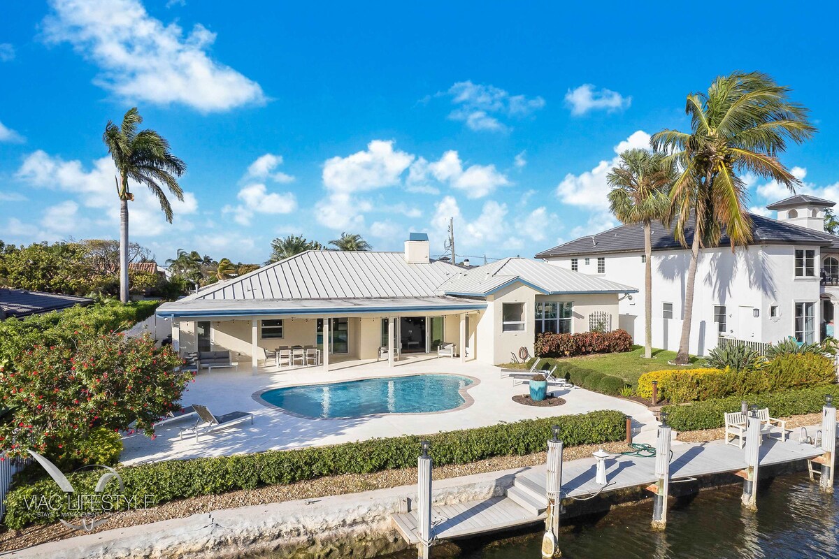 An inviting exterior view of the villa showcases a spacious poolside area surrounded by lush greenery and palm trees. The modern architectural design is complemented by large windows and a sleek metal roof, offering scenic waterfront access and a serene outdoor setting.
