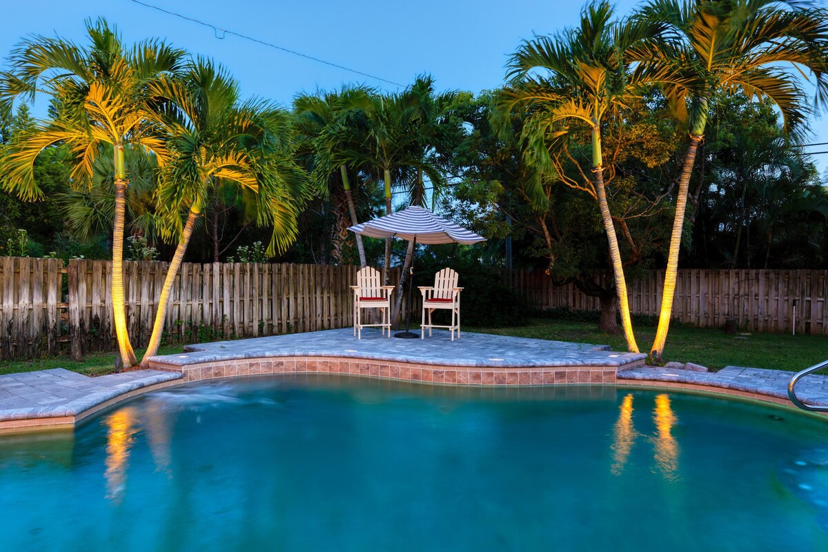 An inviting pool area is surrounded by lush palm trees, softly illuminated by lights. Two adirondack chairs sit under a shaded canopy, offering a serene spot for relaxation. The gentle glow reflects on the water's surface, creating a tranquil evening setting.
