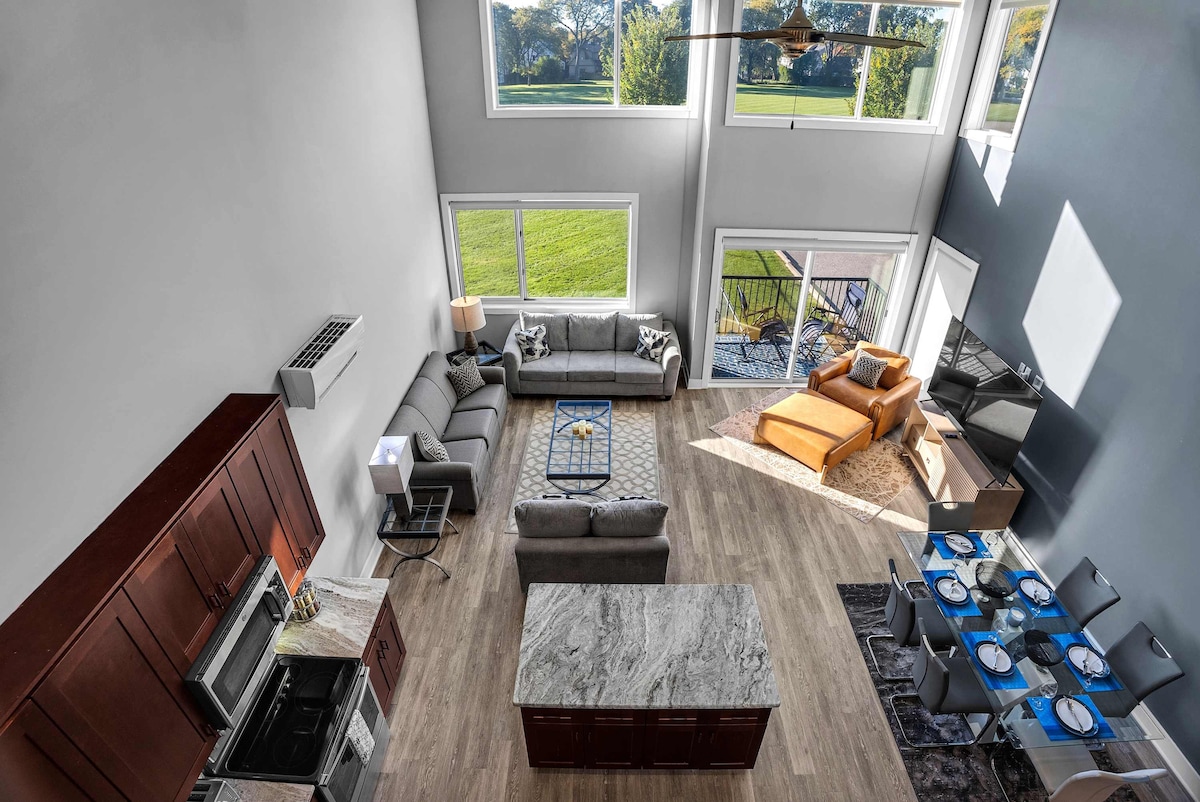 A spacious open-concept living area is highlighted by large windows offering views of green parkland. Comfortable seating is arranged around a coffee table. A modern kitchen with a large island and high-end appliances is visible along with a dining area set for six.