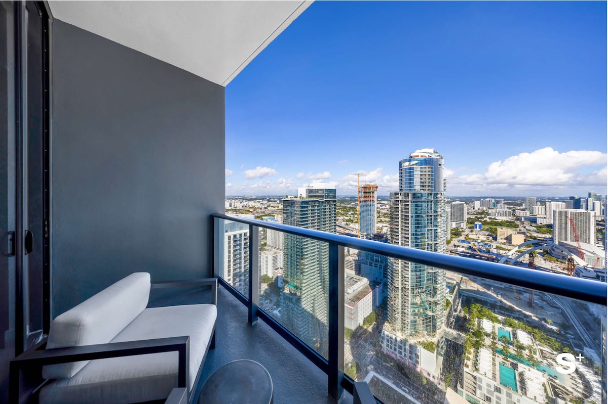 A private balcony offers stunning views of the city skyline and surrounding architecture. A modern, light-colored sofa and a small table provide a comfortable space to relax and enjoy the scenery against a backdrop of clear blue skies.