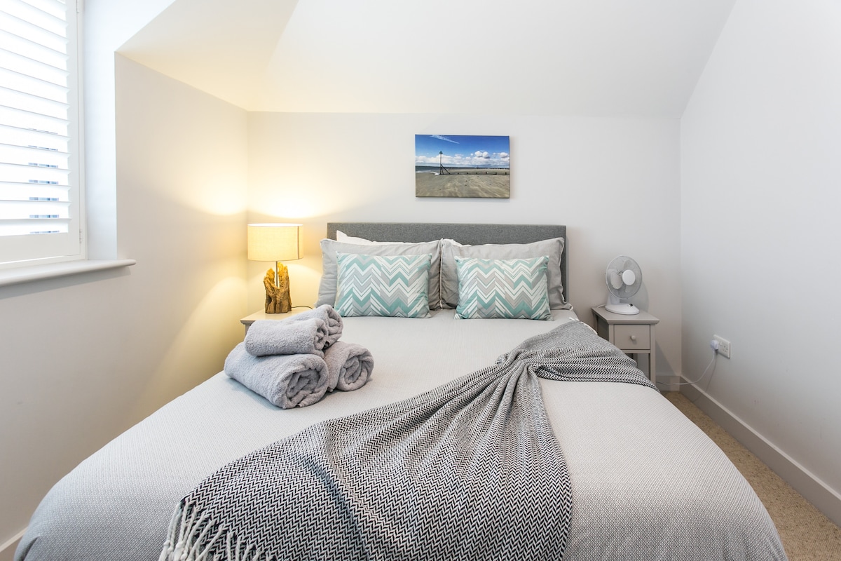 A well-appointed bedroom is presented with a spacious bed adorned with decorative pillows and a soft throw blanket. A bedside table with a lamp and a fan are included, along with a coastal artwork above the bed, contributing to a serene atmosphere.