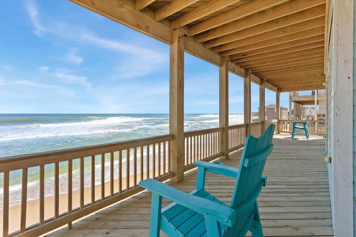 A furnished deck offers a tranquil space with blue Adirondack chairs, providing a perfect spot to enjoy views of the ocean and sandy beach. Waves can be seen gently rolling onto the shore under a clear blue sky.