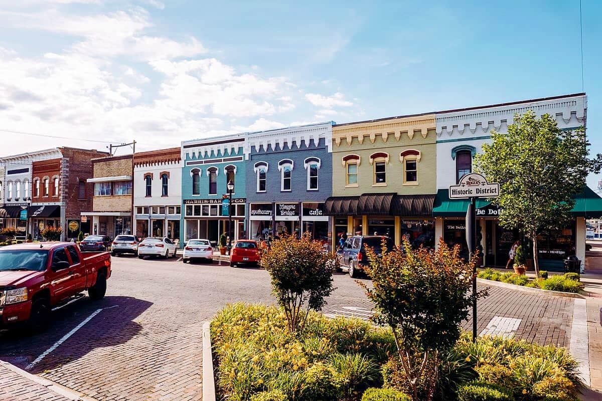 A charming street scene is depicted with colorful historic buildings lining the block. Grass and low shrubbery are visible in the foreground, along with parked vehicles. The sky features soft clouds, suggesting a pleasant day in a vibrant district that invites exploration.