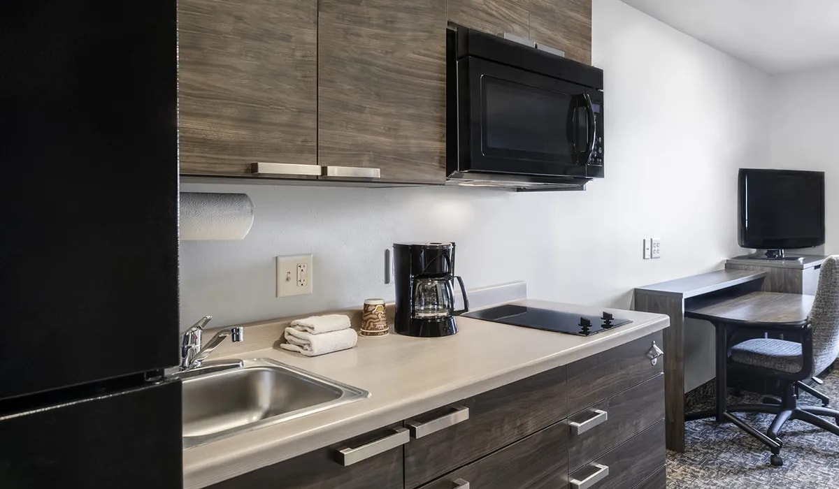 A compact kitchen area features a sleek countertop with a sink, coffee maker, and a microwave. The cabinetry is finished in a warm wood tone. An office workspace with a chair is positioned nearby, and a flat-screen television is mounted on the wall.