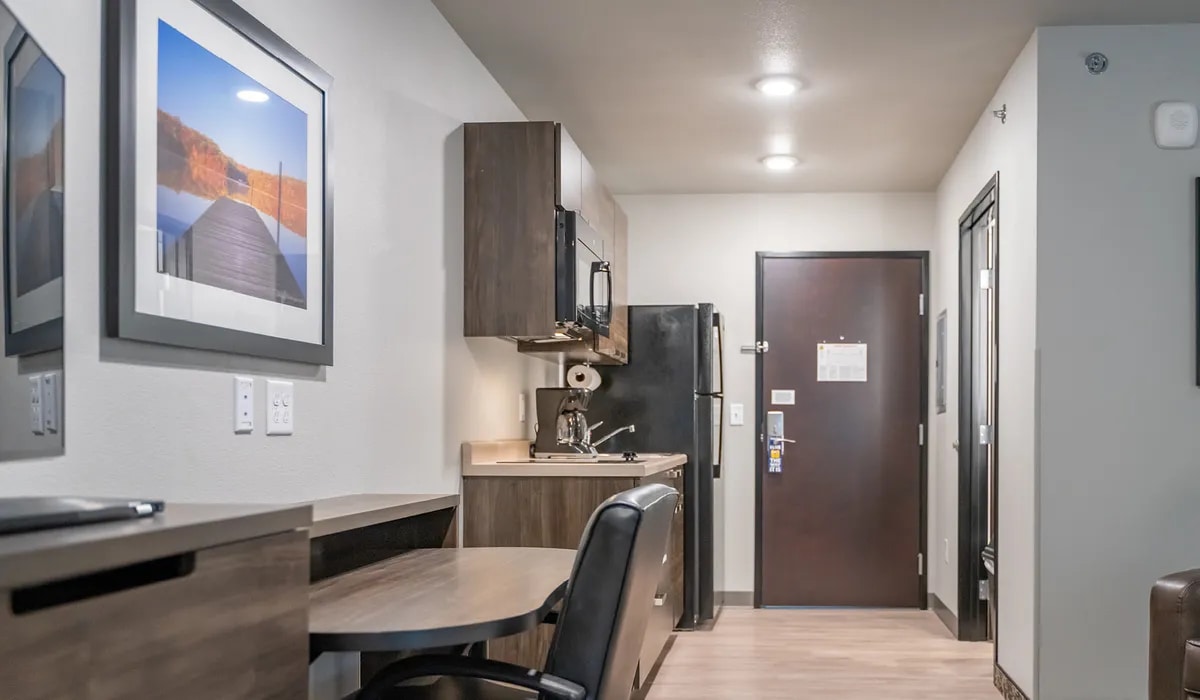 A well-organized hotel room features a kitchenette with a refrigerator, microwave, and coffee maker. A small dining table is positioned beside a workspace. A dark wood door leads to the guest room, while artwork depicting a serene landscape is displayed on the wall.