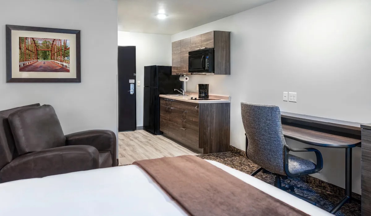 The cozy room features a queen bed with a recliner and desk nearby. A kitchenette equipped with a microwave and mini fridge is visible. Light wood tones enhance the modern decor. A framed landscape print is displayed on the wall, adding a touch of color.