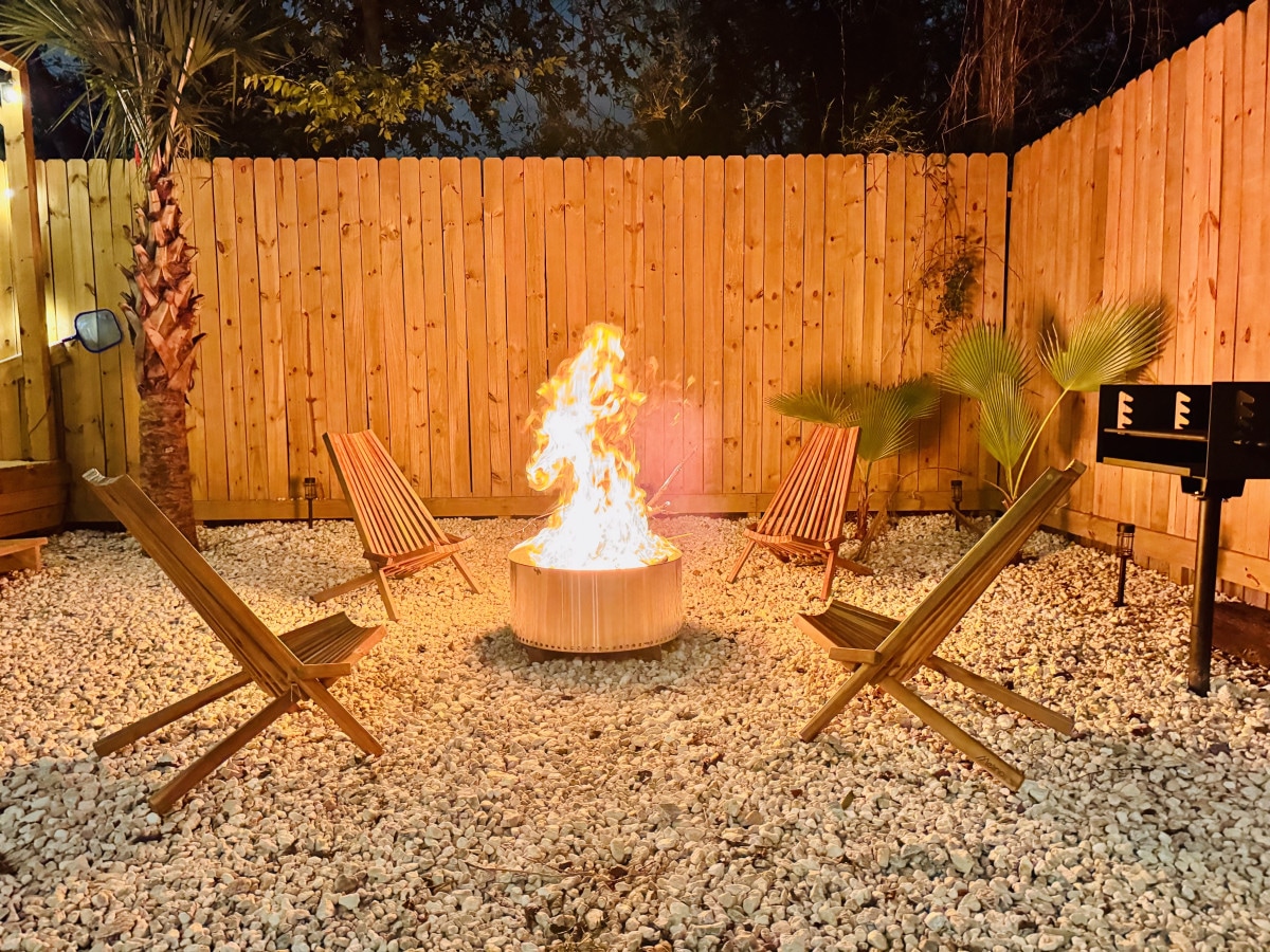 A cozy fire pit surrounded by four wooden chairs is set on pebbled ground, creating a warm gathering spot. The area is enclosed by a wooden fence, with palm trees and greenery adding a touch of nature to the evening ambiance.