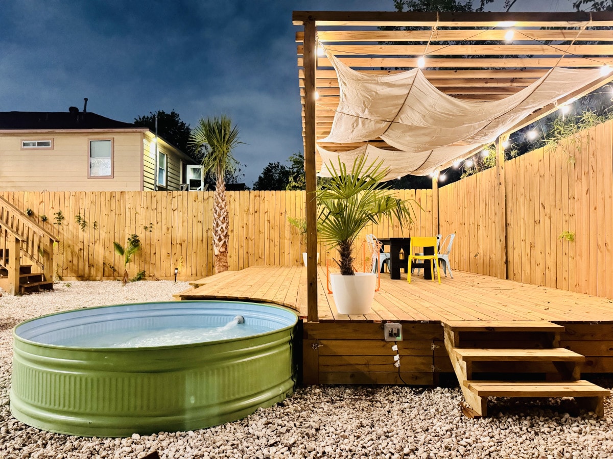 A spacious wooden deck is featured with a stock tank pool, surrounded by gravel and greenery. A large fabric canopy offers shade, and a dining table with chairs is visible, creating an inviting outdoor space for relaxation and meals.