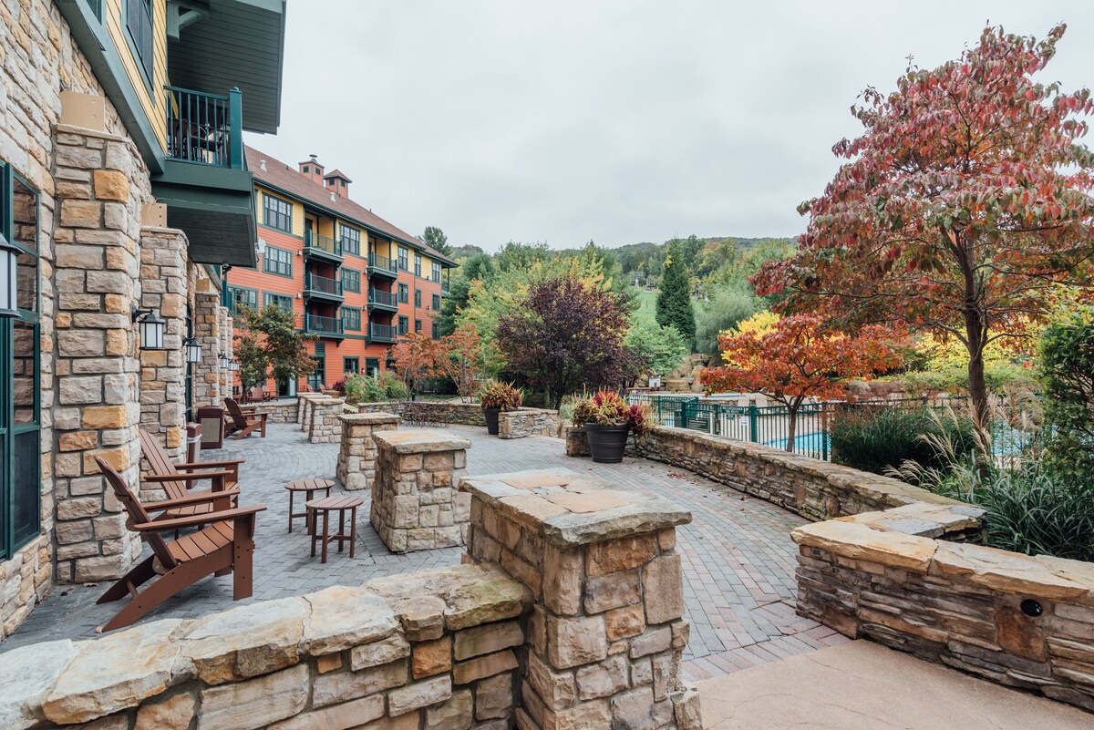 An outdoor patio area is featured, surrounded by stone walls and complemented by rustic wooden chairs. Seasonal trees add color to the landscape, while a serene pool is visible in the background, contributing to a relaxed ambiance.
