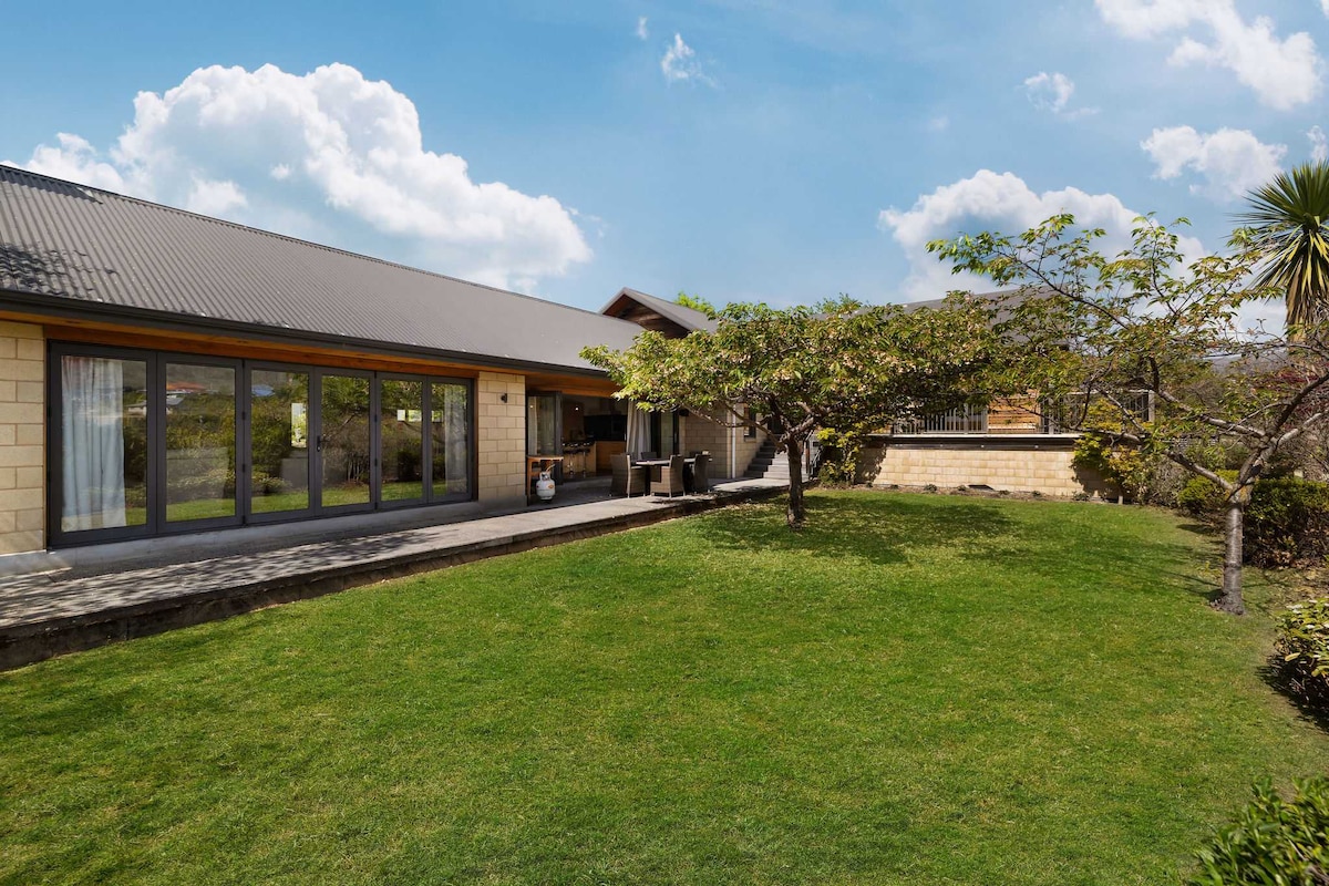 A well-maintained garden is seen, with a lush green lawn and a flowering tree providing shade. Glass sliding doors lead to a spacious outdoor patio area, creating a seamless connection between the interior and the landscaped surroundings.