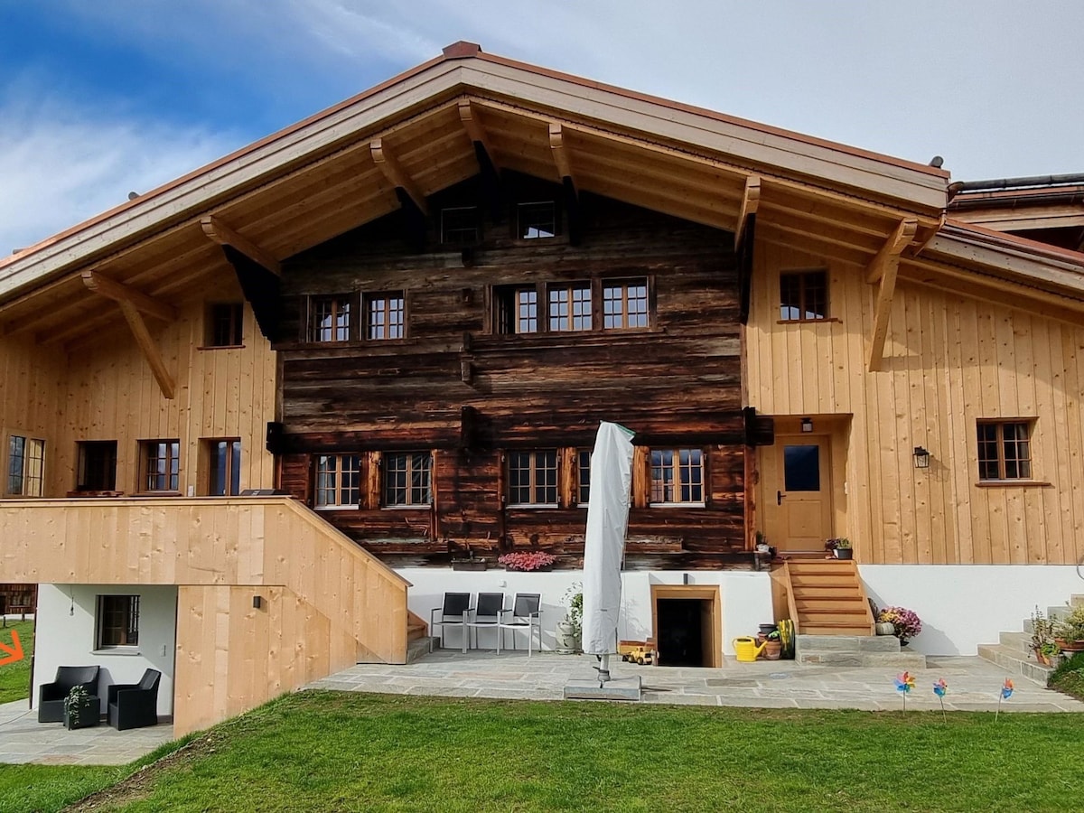 The exterior of a charming, renovated farmhouse displays a combination of traditional wooden architecture and modern elements. A spacious terrace is visible, furnished with outdoor seating, alongside a well-kept lawn and flower beds. Large windows offer views into the building, enhancing the inviting atmosphere.