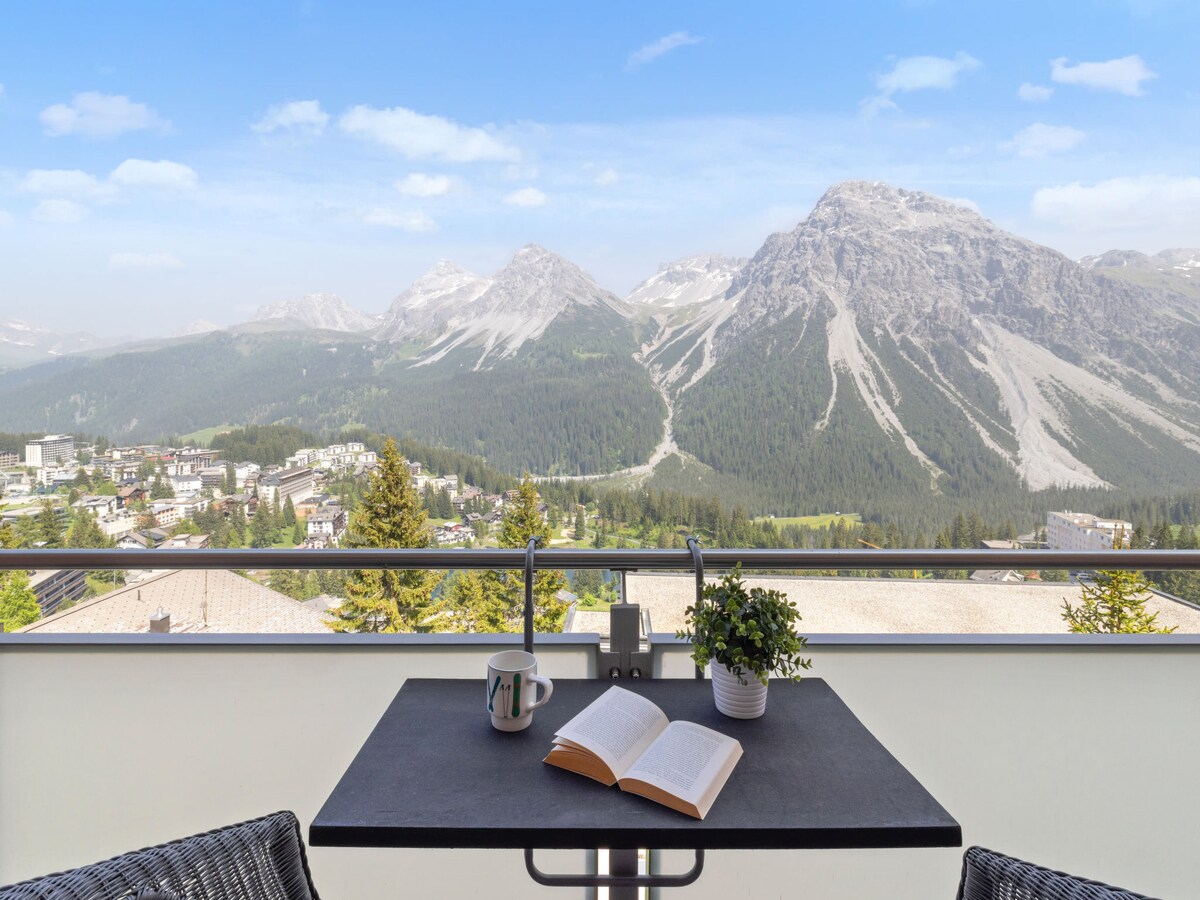 A small balcony is furnished with a table and two chairs, providing a space to relax. An open book and a coffee cup are placed on the table. The panoramic view showcases mountains and the town below, under a clear blue sky.