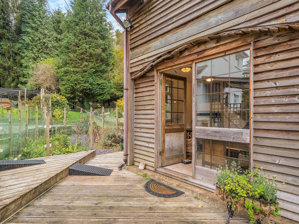 An entrance to the lodge is framed by a wooden deck, leading to large glass doors that invite natural light into the interior. Surrounding greenery is visible, with a garden area beyond the entrance, enhancing the connection to nature.