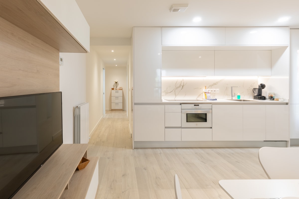 A modern kitchen is displayed, featuring sleek white cabinetry and an integrated oven. Soft lighting highlights the clean surfaces, and small appliances are neatly arranged. A living area with a television is visible, creating a harmonious flow in this open-concept space.