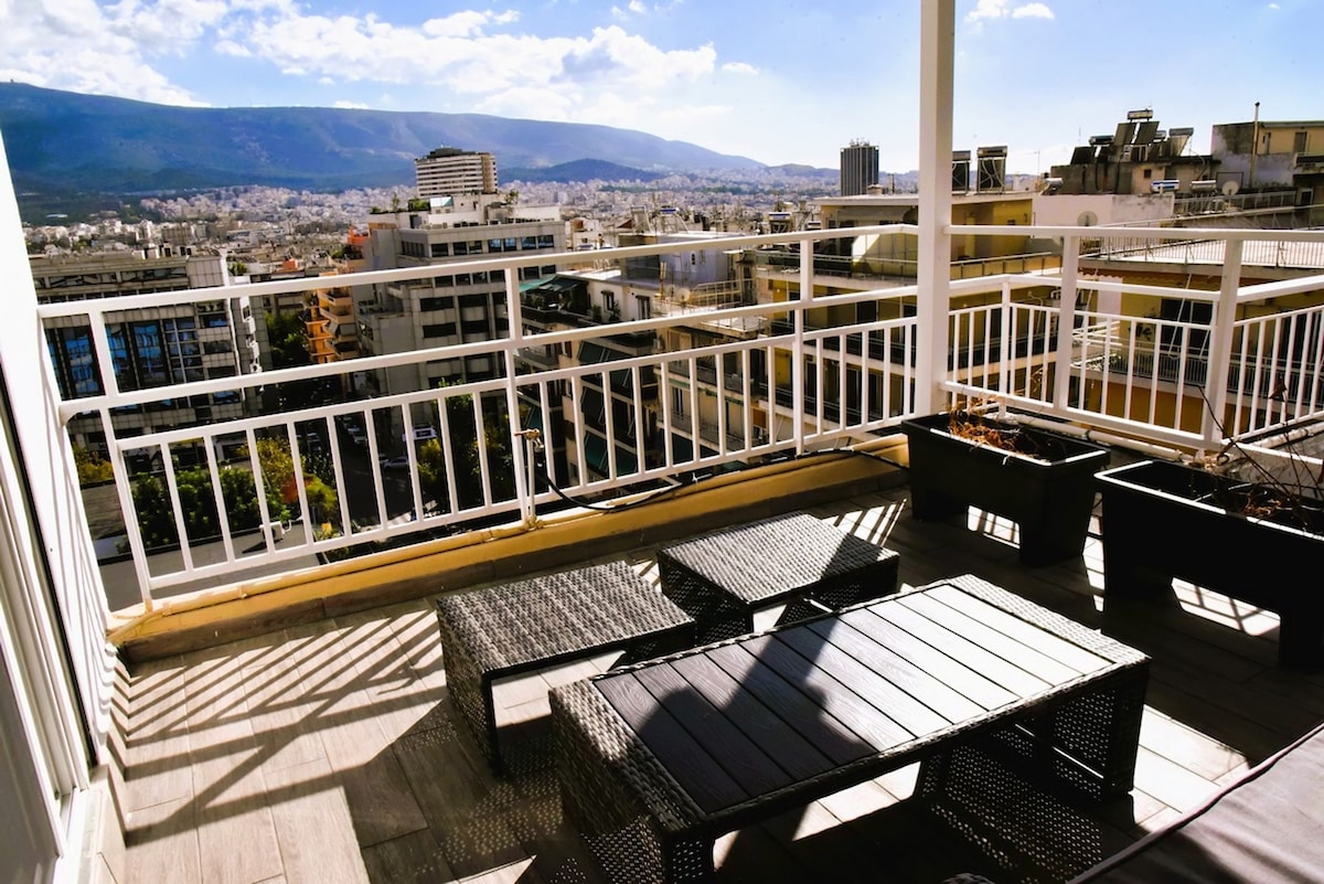 A sunlit balcony features outdoor furnishings including a dark wicker seating area and a low table. Planters are arranged along the railing, with a backdrop of distant mountains and cityscape visible under a clear blue sky.