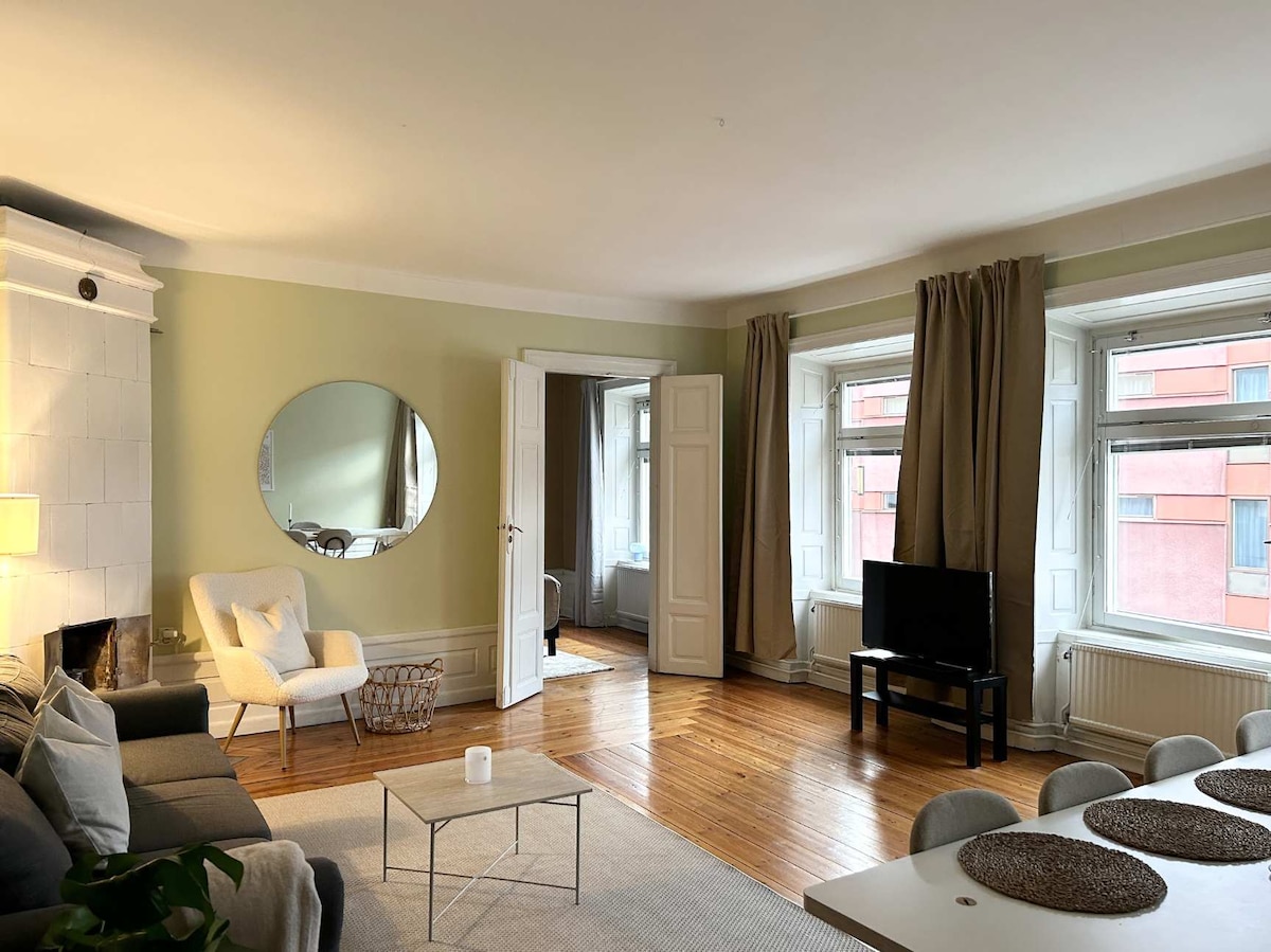A welcoming living area is presented, featuring a comfortable sofa and a round coffee table. Large windows allow natural light to fill the space, while soft curtains frame the views. A wall mirror enhances the sense of openness, and a television is positioned for casual viewing.