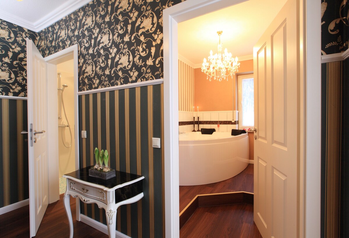 An elegantly designed interior displays an inviting bathroom featuring a luxurious whirlpool tub. Decorative wallpaper with floral patterns enhances the ambiance, while a stylish console table with a plant decorates the adjacent hallway. Soft lighting creates a warm atmosphere throughout the space.