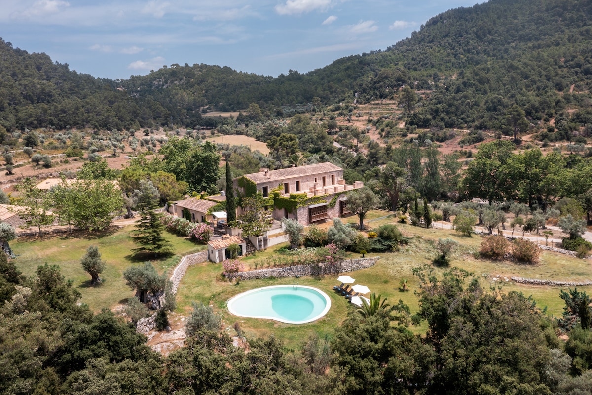 An expansive villa nestled in lush greenery, surrounded by rolling hills. A private swimming pool is visible in the foreground, with sun loungers positioned nearby. The landscape features a variety of trees and garden spaces, creating a serene outdoor environment.