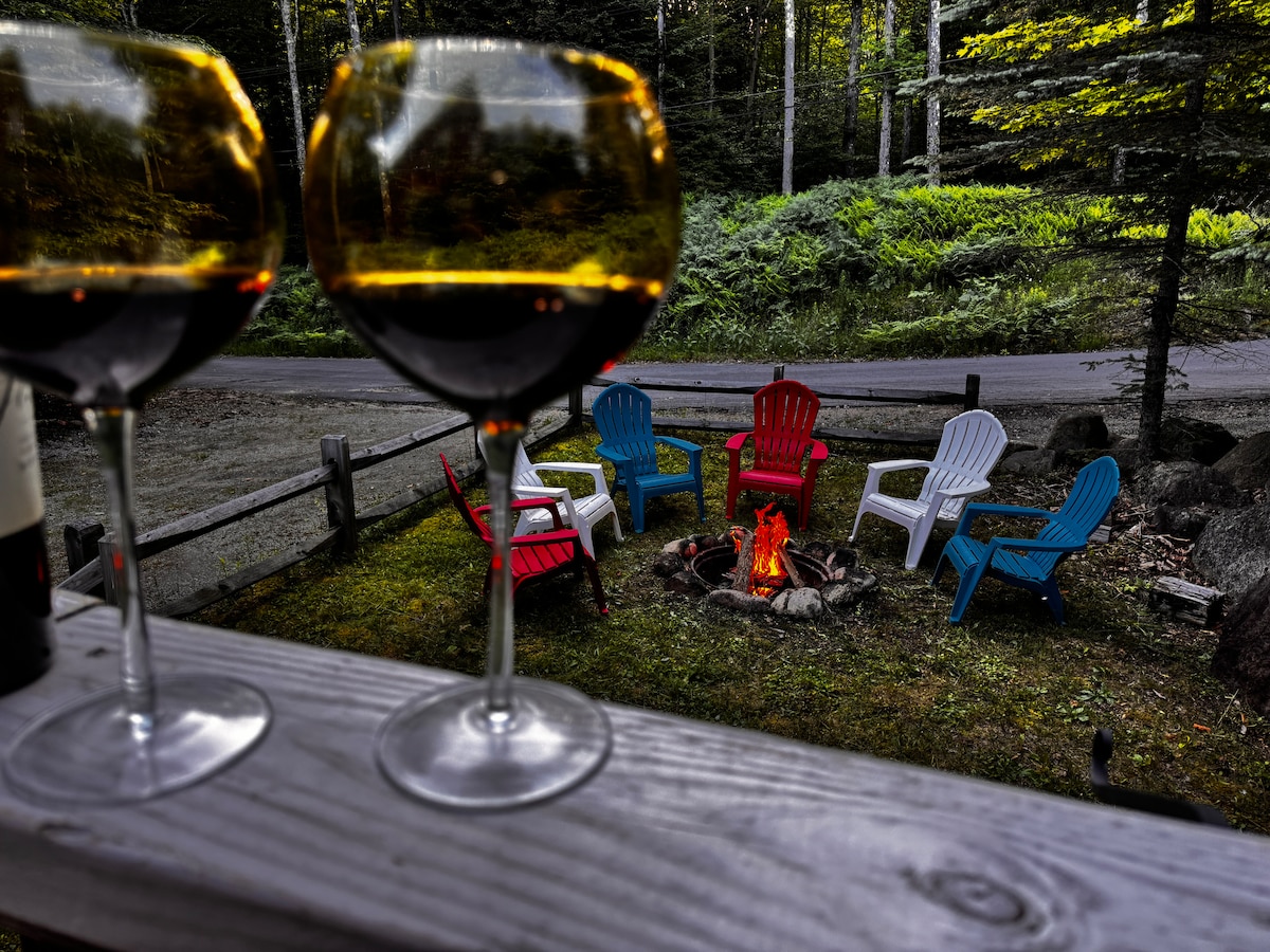 A fire pit is surrounded by brightly colored Adirondack chairs in red, blue, and white. Two glasses filled with dark liquid are in the foreground, set on a wooden railing. Lush greenery and a road are visible in the background, contributing to a peaceful outdoor setting.